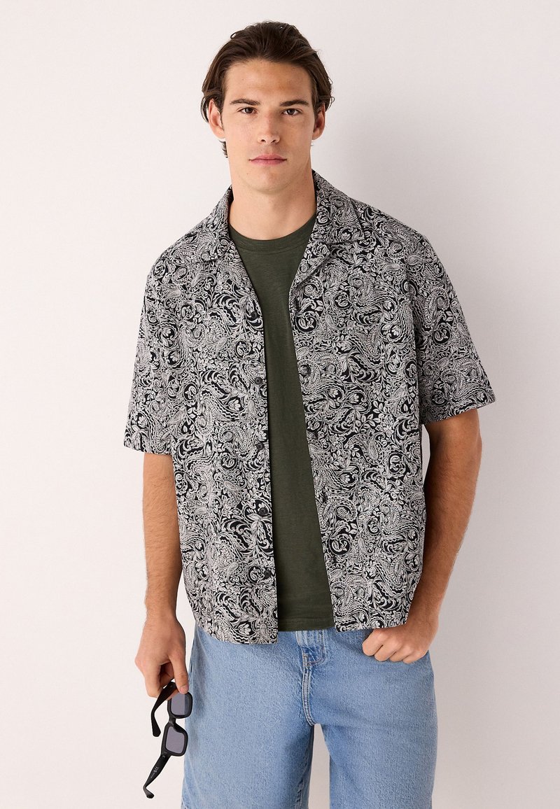 Camicia a maniche corte con motivo paisley bianco e nero, tessuto leggero, collo aperto, indossata sopra una maglia verde, abbinata a jeans azzurri.