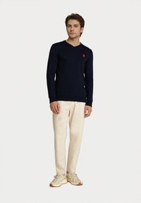 Hombre de pie vistiendo un suéter de pico navy con logo rojo, pantalones crema y zapatillas beige sobre un fondo blanco.