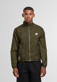 Muž v olivově zelené zipové bundě s malým bílým logem na hrudi a černých kalhotách na jednobarevném pozadí.