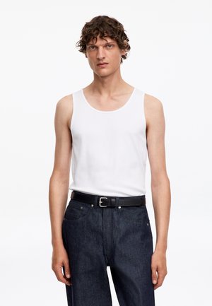 Uomo con capelli ricci che indossa una canotta bianca senza maniche infilata in jeans blu scuro con una cintura nera, in piedi davanti a uno sfondo bianco.