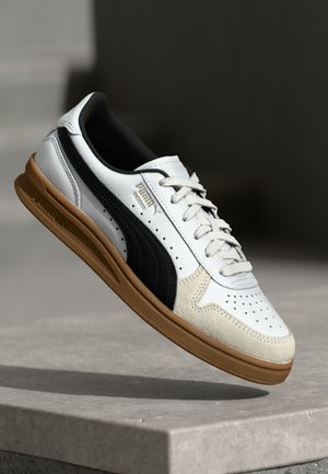 Baskets d’intérieur blanches Puma avec bande noire, bout en daim beige, semelle en gomme et lacets blancs flottant au-dessus d’une surface en béton.