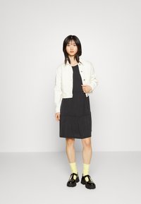 Casaco de denim branco sobre um vestido preto com camadas. Modelo usa meias amarelas e sapatos de plataforma pretos com detalhes amarelos. Design simples e moderno.