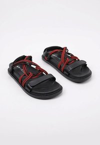 Sandalias negras con detalles de cuerda roja. Presentan una suela plana, dos correas anchas y una superficie texturizada. Diseño moderno con hardware mínimo.