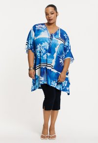 Blauwe bloemen tuniek met korte mouwen, voorzien van een V-hals en decoratieve strikjes. Gepaarde met zwarte capri-broek en sandalen.