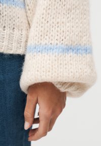 Κοντινό πλάνο ενός χεριού που κρέμεται χαλαρά στο πλάι, φορώντας ένα κρεμ πλεκτό πουλόβερ με ανοιχτό μπλε ρίγα και μπλε τζιν παντελόνι.