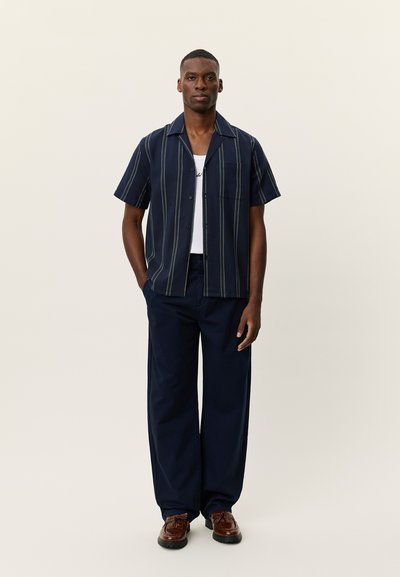 Chemise à manches courtes boutonnée bleu marine avec rayures verticales, associée à un pantalon large bleu marine et des chaussures marron, posant devant un fond neutre.