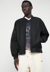 Veste bomber noire avec une texture lisse, fermeture éclair à l'avant et poignets élastiques, portée sur un T-shirt en maille foncée et associée à un pantalon clair.