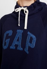 Sudadera azul marino con collarín, que presenta un cordón blanco. Grande, brillante logo azul "GAP" en un estilo de letra audaz en la parte frontal.