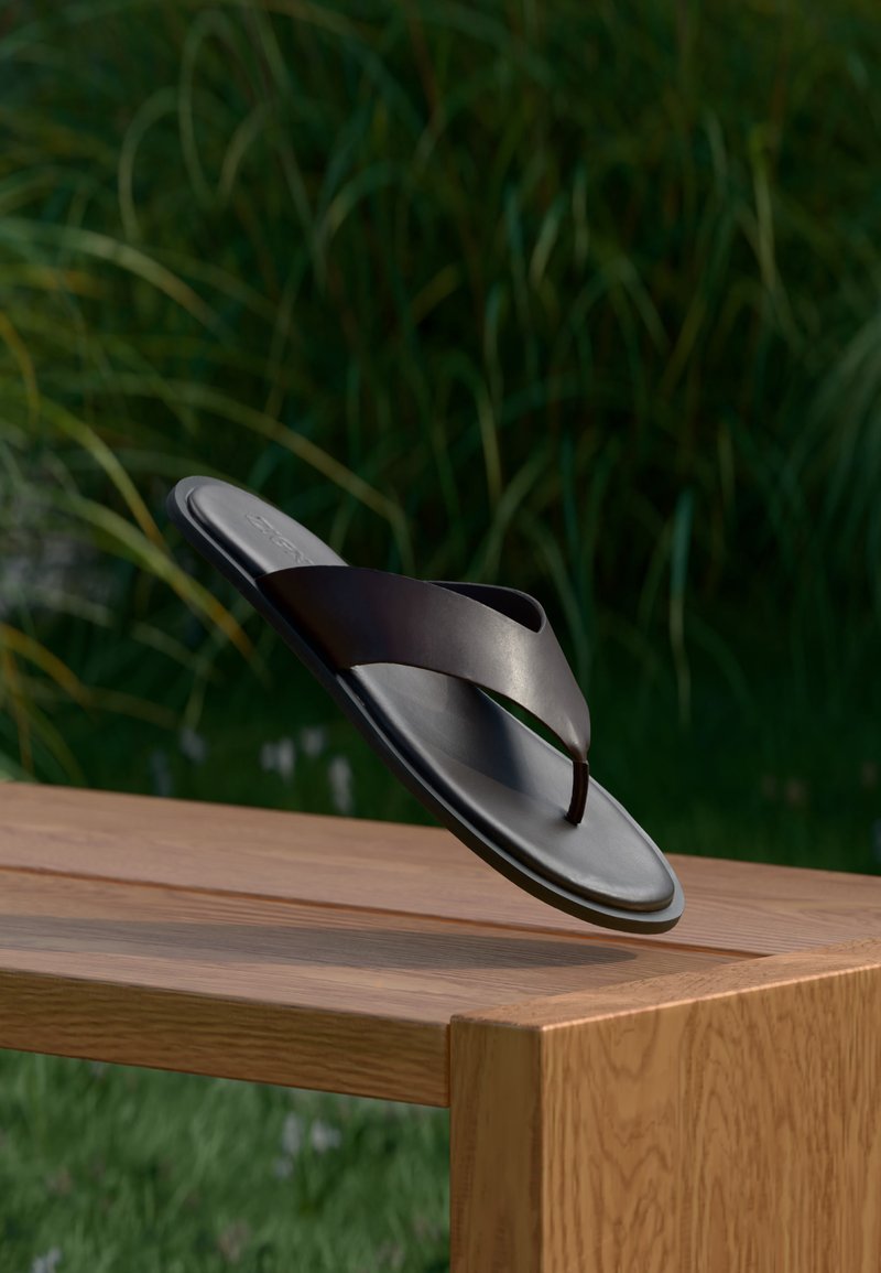 Μαύρο δερμάτινο σανδάλι με λουράκι να αιωρείται πάνω από ένα ξύλινο παγκάκι με πράσινα φύλλα στο θολό φόντο.