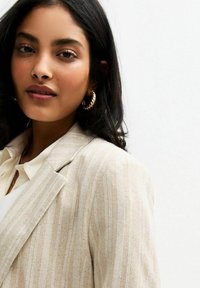 Beige randig blazer i texturerat tyg, med slagkrage, ihop med en vit skjorta och gyllene hoopsringar.