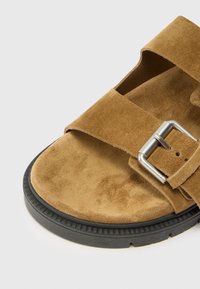 Sandale din suede maro cu două curele late, o cataramă metalică și o talpă din cauciuc negru cu un model texturat.