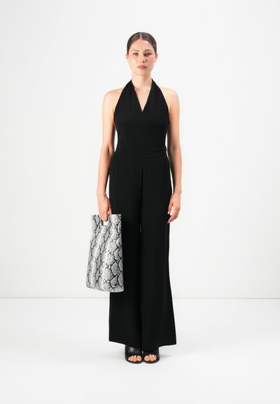 Woman in black halter jumpsuit holds a large gray snakeskin-patterned tote, standing against a plain white background.