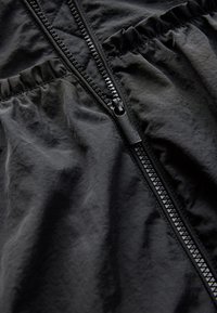 Black, lightweight jacket featuring a prominent zipper, gathered texture along the edges, and a smooth, matte finish.