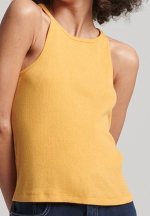 Person iført en ærmeløs gul ribstrikket tanktop og mørkeblå jeans mod en neutral baggrund.
