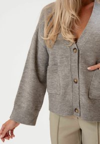 Szürke kötött kardigán V-nyakkal, hosszú ujjakkal és két elülső zsebbel. Barna gombokkal és puha textúrával rendelkezik.