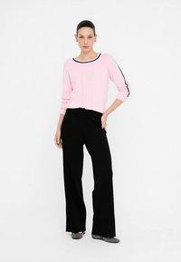 Maglione rosa chiaro lavorato a maglia con rifiniture nere, abbinato a pantaloni neri a gamba larga. La modella è in piedi su uno sfondo bianco, indossando scarpe basse grigie.