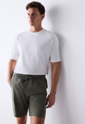 Mann trägt ein weißes kurzärmeliges Hemd und olivgrüne Kordelzug-Shorts und steht mit den Händen in den Taschen an einer weißen Wand.