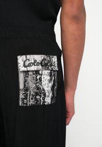 Μαύρο αθλητικό σορτς με υφασμάτινο υφήμα σε ασπρόμαυρο μοτίβο και το κείμενο «Cala Cala» στην πίσω τσέπη. Μαλακό ύφασμα.