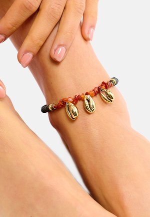 Cheville perlée avec des pierres orange et marron, décorée de coquillages dorés de type cauris et d'accents noirs, portée sur une cheville couleur chair.