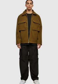 Veste en polaire marron avec un col, deux poches avant, fermeture éclair, portée sur une chemise noire, assortie à un pantalon cargo noir ample et des baskets blanches.
