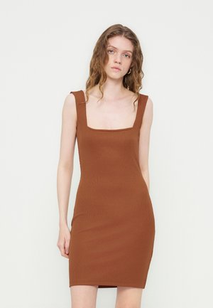 Ung kvinne med bølgete hår har på seg en brun, ermeløs, tettsittende kjole med brede stropper og firkantet utringning mot en ensfarget lys bakgrunn.