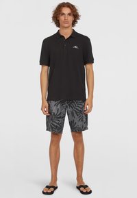 Sort polo med krave og logo på brystet, parret med grå shorts med et sort bladmønster. Sandaler fuldender looket.