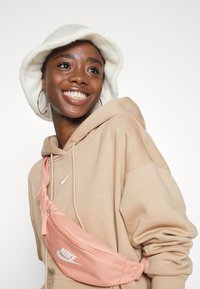Nike fanny pack šviesiai rožinės spalvos su logotipu, dėvima ant šviesiai smėlinio spalvos džemperio su gobtuvu. Asmuo dėvi baltą pūkuotą kibirėlį ir auskarus žiedus. Lygus paviršius.