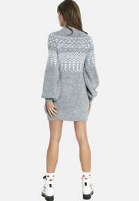 Kobieta nosząca szarą dzianinową sukienkę z białym wzorem, z bufkami na rękawach, białe skarpetki z czerwonymi gwiazdkami oraz białe sznurowane buty, odwrócona tyłem.