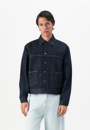 Mann mit dunklen Haaren, der eine dunkelblaue Jeansjacke mit Fronttaschen und hellblauen Jeans trägt, stehend vor einem weißen Hintergrund.