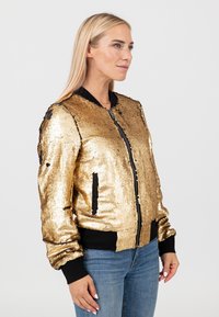 Donna con lunghi capelli biondi che indossa una giacca bomber dorata con paillettes, polsini neri e jeans blu, in piedi davanti a uno sfondo chiaro e semplice.