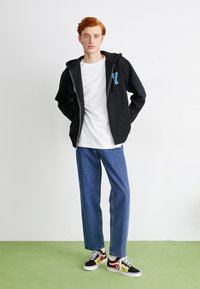 Schwarzer Zip-Hoodie mit blauem "Y"-Logo, weißem T-Shirt, blauen Jeans und mehrfarbigen Sneakers mit roten, gelben und schwarzen Akzenten auf grünem Boden.