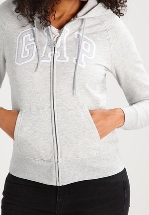Frau trägt hellgrauen Kapuzenpullover mit Reißverschluss und GAP-Logo, Hände in den vorderen Taschen, kombiniert mit schwarzen Hosen.