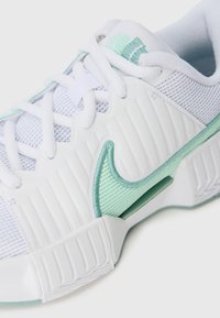 Zapatilla blanca con el swoosh de Nike en verde claro, parte superior de malla, suela gruesa y acanalada, y cordones blancos sobre un fondo blanco.