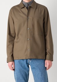 Chaqueta marrón de botones con cuello y un bolsillo en el pecho, hecha de una tela suave. Combinada con jeans azul claro.