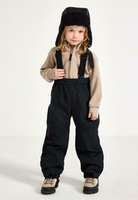 Beige fleece zip-up-tröja och svarta vattenavvisande byxor med hängslen. Barnet bär en svart pälsfoderhatt och beige stövlar med snörning.