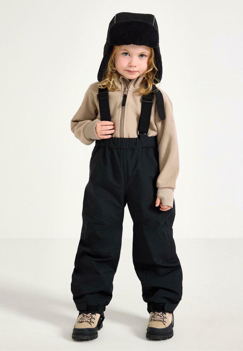 Beige fleece zip-up-tröja och svarta vattenavvisande byxor med hängslen. Barnet bär en svart pälsfoderhatt och beige stövlar med snörning.