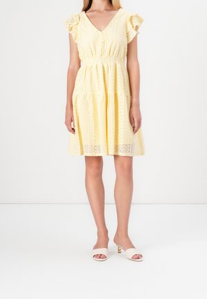 Żółta sukienka z koronkowym wzorem, dekoltem w serek, falbankami na rękawach i elastyczną talią. Do niej nude klapki na obcasie z odkrytymi palcami.