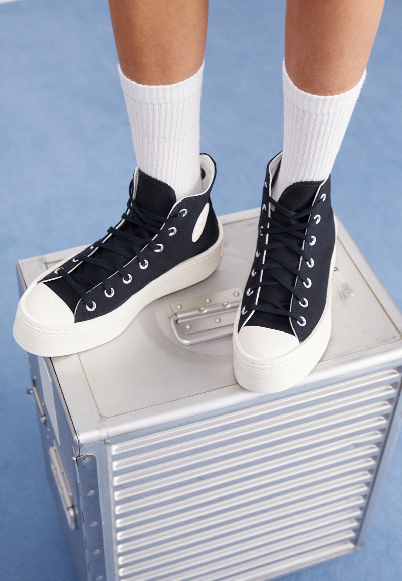 Zapatillas altas negras con punteras y cordones de goma blanca, apoyadas sobre una caja metálica, combinadas con calcetas blancas de canalé.