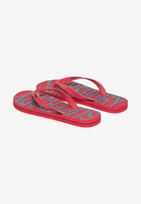 Des tongs en caoutchouc rouge avec une semelle texturée et une marque grise. Les brides sont également rouges, présentant un motif en zigzag pour une meilleure adhérence.