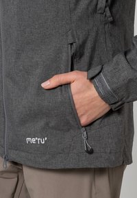 Grå utomhusjacka med texturerat tyg, ficka med dragkedja och justerbar dragsko i botten. Logotyp från märket synlig på framsidan.