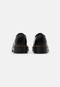 Paire de chaussures en cuir noir avec semelles épaisses rainurées et couture jaune, vues de dos sur un fond blanc.