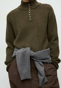 Suéter de punto verde oliva con cuello alto, que cuenta con una tapeta de botones con detalles plateados, combinado con un suéter de punto gris atado en la cintura.