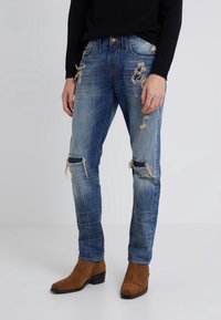 Blå denimjeans med slitna detaljer, inklusive hål och fransade kanter. Åtsittande design i kombination med bruna mockastövlar.