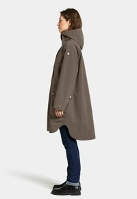 Veste imperméable marron avec capuche, poches latérales et accents de boutons-pression. Présente une coupe décontractée et un ourlet arrondi, associée à un jean foncé et des bottes.