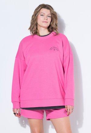 Kobieta ubrana w jaskraworóżową oversize bluzę i pasujące szorty z czarnym napisem i grafiką serca, stojąca przy białej ścianie.