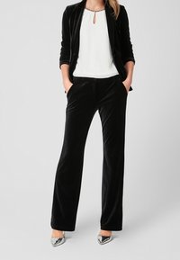 Musta samettinen bleiseri ja housut yhdistettynä valkoiseen, avainreikäyksityiskohtaa sisältävään topiin sekä hopeisiin, kärjestä teräviin kenkiin. Sileä pinta, räätälöity istuvuus.