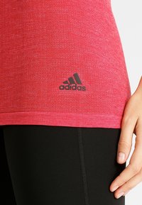 Červený sportovní top s texturovaným povrchem, s černým logem Adidas poblíž spodního lemu. Top je spárován s černými legínami.