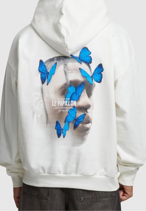 Sweat à capuche blanc avec un graphisme au dos représentant le visage d’un homme partiellement recouvert par sept papillons bleus vibrants et le texte « Le Papillon ».