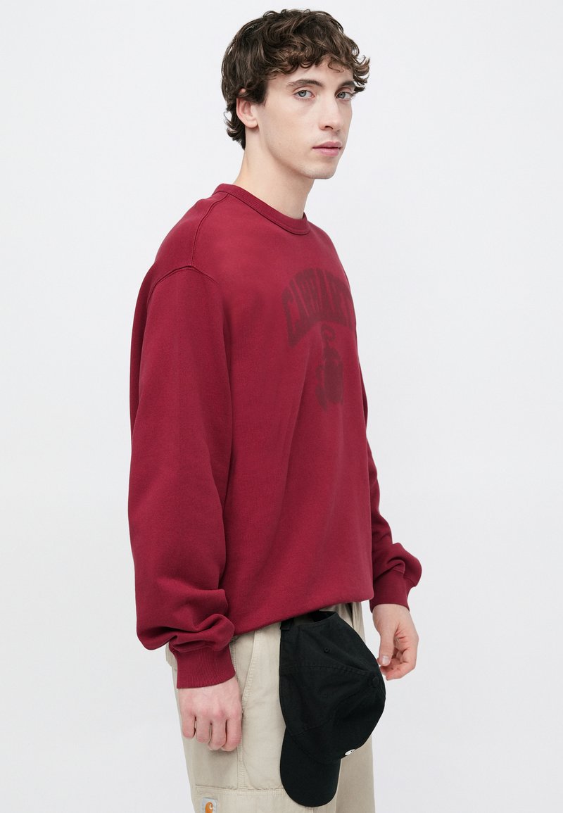 Sweat-shirt bordeaux à col rond et manches longues, avec un imprimé graphique subtil. Associé à un pantalon cargo clair et une casquette noire.