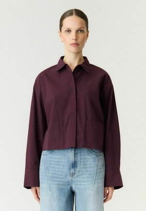 Femme à la peau claire et aux cheveux bruns attachés porte une chemise à manches longues violet foncé et un jean taille haute bleu clair, debout devant un fond uni.
